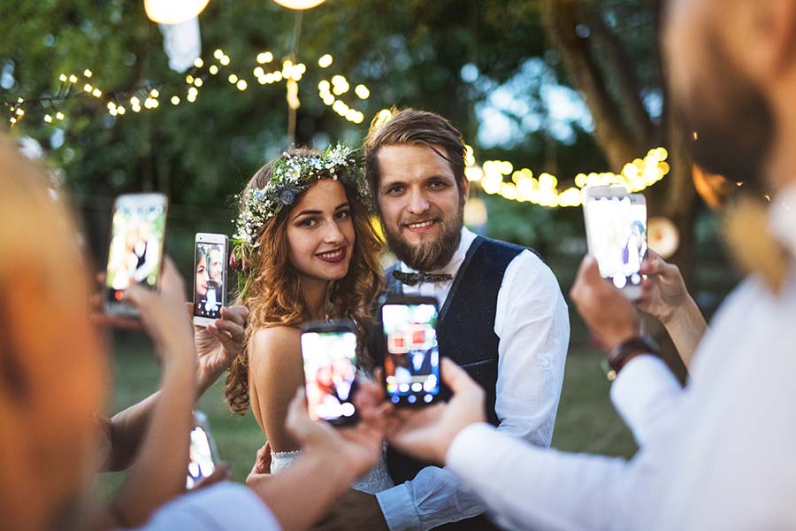 Guests with smartphones taking photo of bride and groom at wedding reception outside.