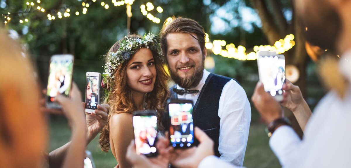 Guests with smartphones taking photo of bride and groom at wedding reception outside.